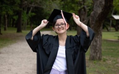 A smiling graduate stands outdoors on a gravel path, holding the sides of her graduation cap with both hands. She wears a black cap and gown over a white top and lavender pants. Trees and grass surround the path, creating a peaceful, celebratory scene.