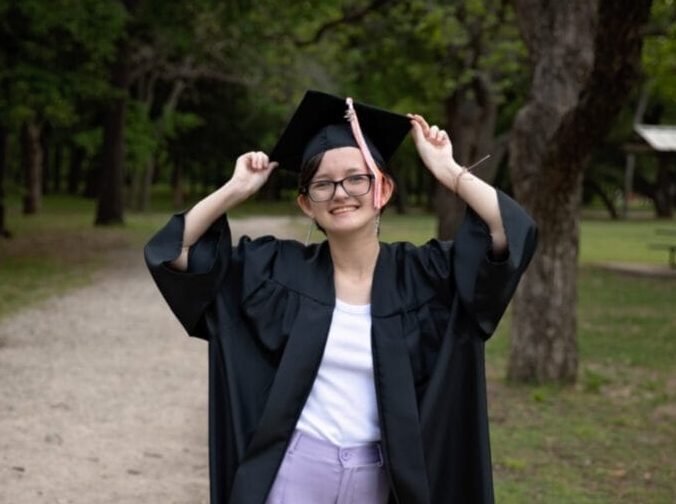 A smiling graduate stands outdoors on a gravel path, holding the sides of her graduation cap with both hands. She wears a black cap and gown over a white top and lavender pants. Trees and grass surround the path, creating a peaceful, celebratory scene.