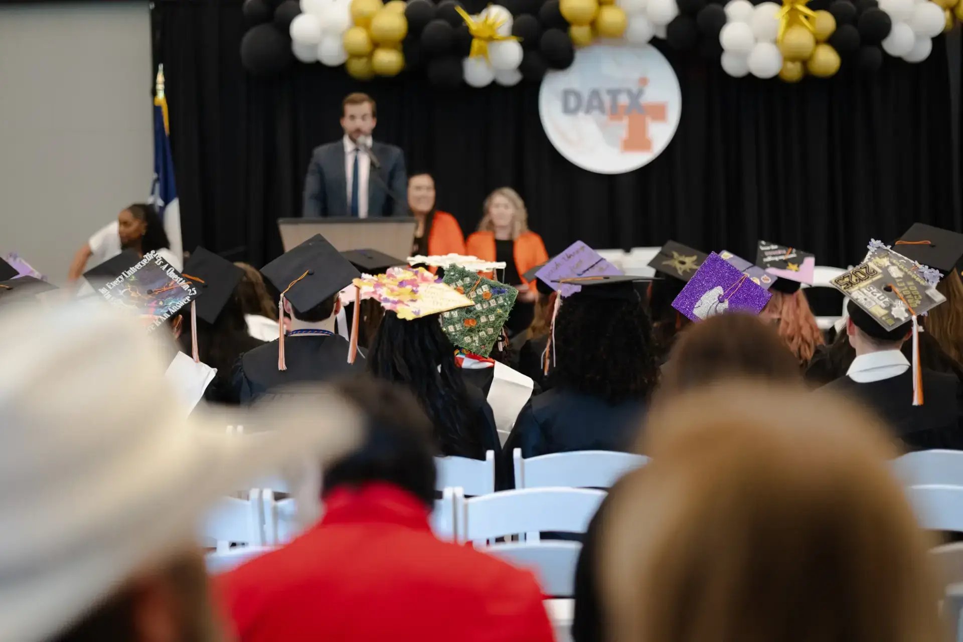 Graduation ceremony with students in decorated caps facing a speaker on stage.