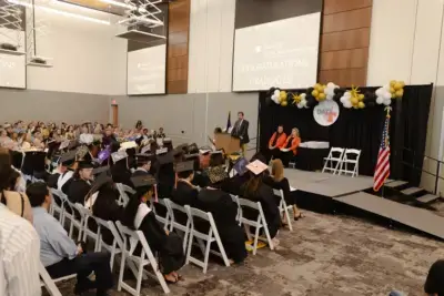 Graduation ceremony in a hall, speaker at a podium addressing graduates in caps and gowns.