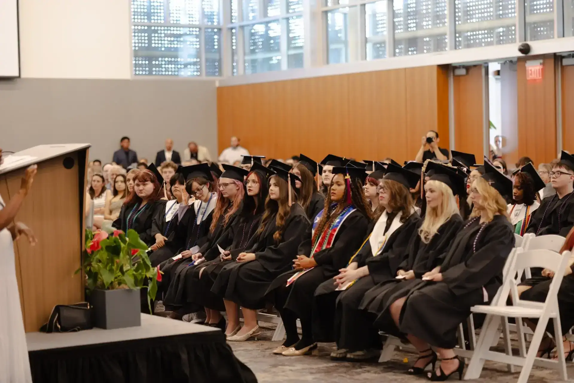 A group of graduates in black gowns and caps sit attentively in rows during a ceremony.