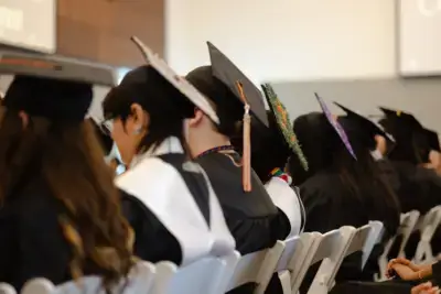 Graduates sit in a row wearing caps and gowns, facing forward.