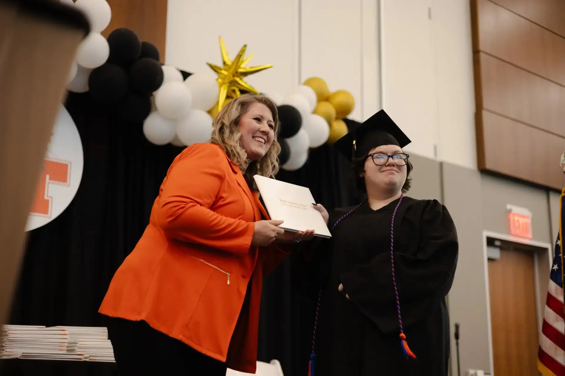 A person in a bright orange blazer smiles while handing a diploma to a graduate in a black cap and gown.