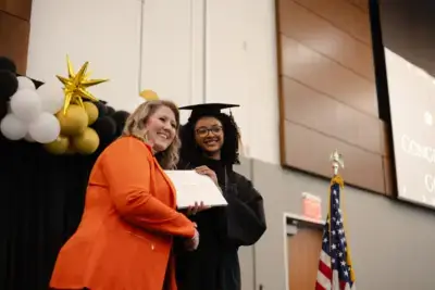 A graduate in a black cap and gown smiles and receives a diploma from a woman in an orange jacket