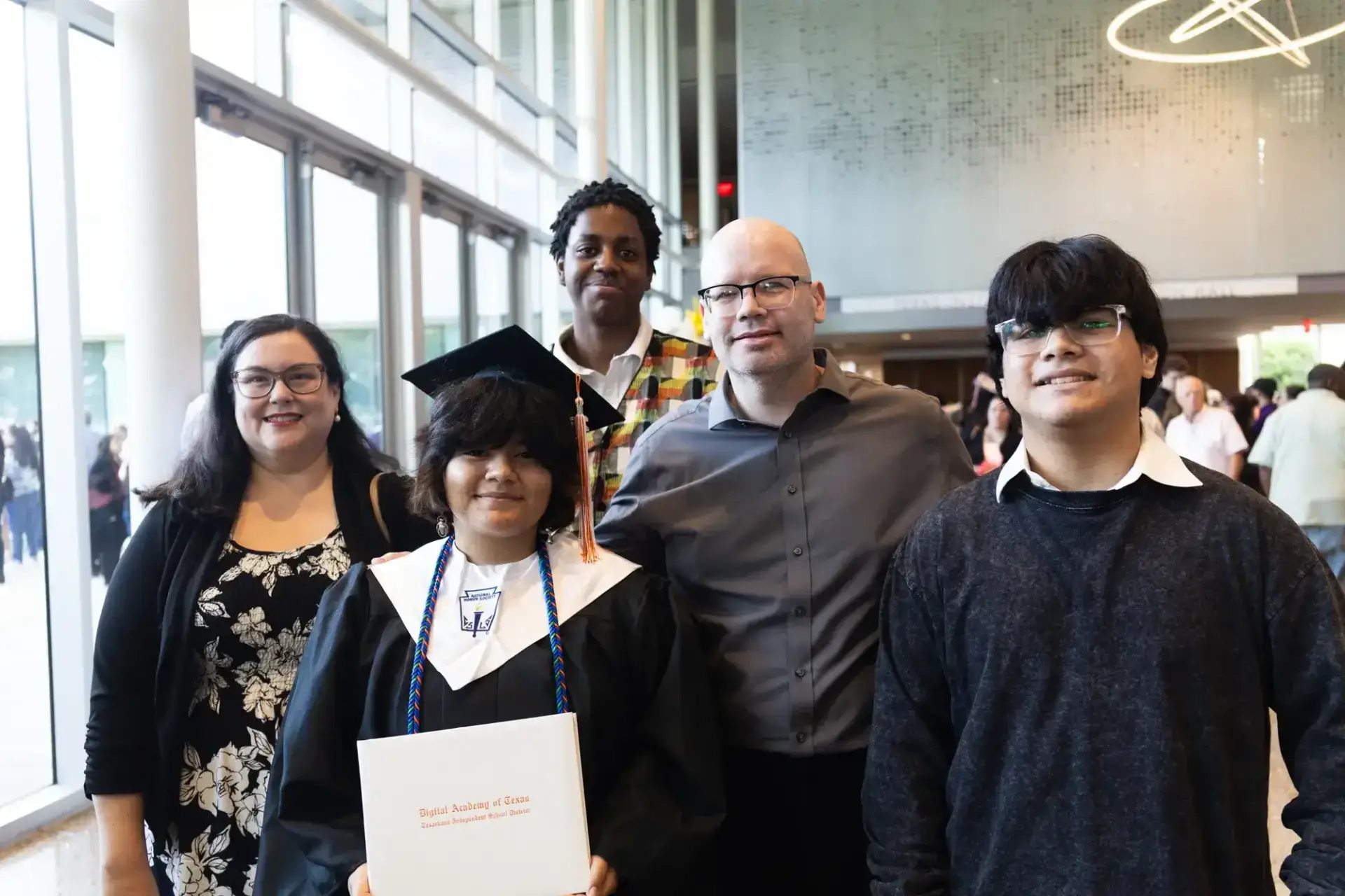 A group photo showing five people indoors at a graduation event. The graduate, wearing a cap and gown, holds a diploma, surrounded by smiling supporters.