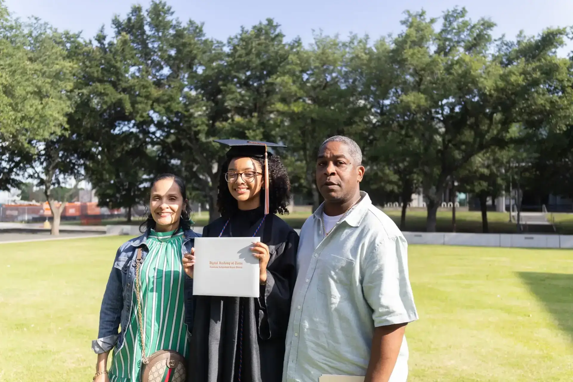 A graduate in cap and gown stands proudly between two smiling individuals, holding a diploma.