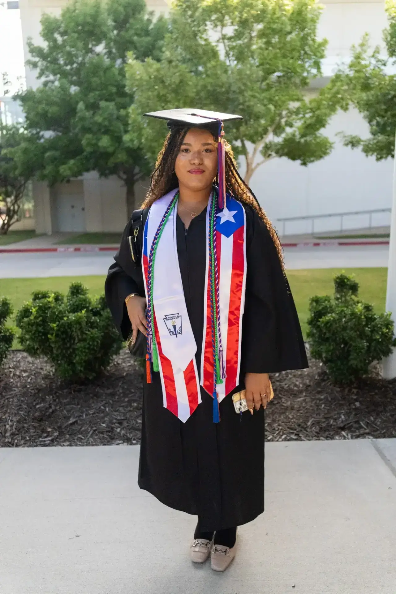 A graduate stands proudly in a black cap and gown, adorned with a Puerto Rican flag sash and honor cords